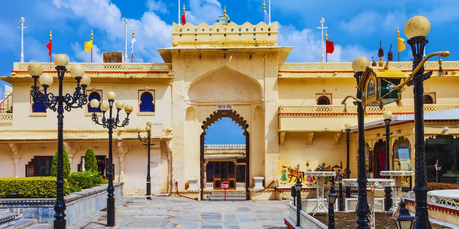 Ornate black lampposts frame the entrance to a pale yellow, historic Indian palace (Udaipur City Palace) structure under a bright blue, cloudy sky.